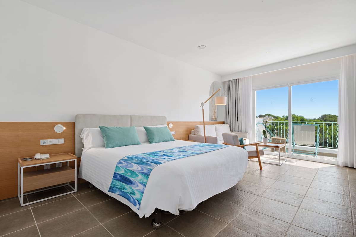 A modern hotel room with a large bed, a bedside table with a lamp, a small table with chairs, and a balcony with a view.