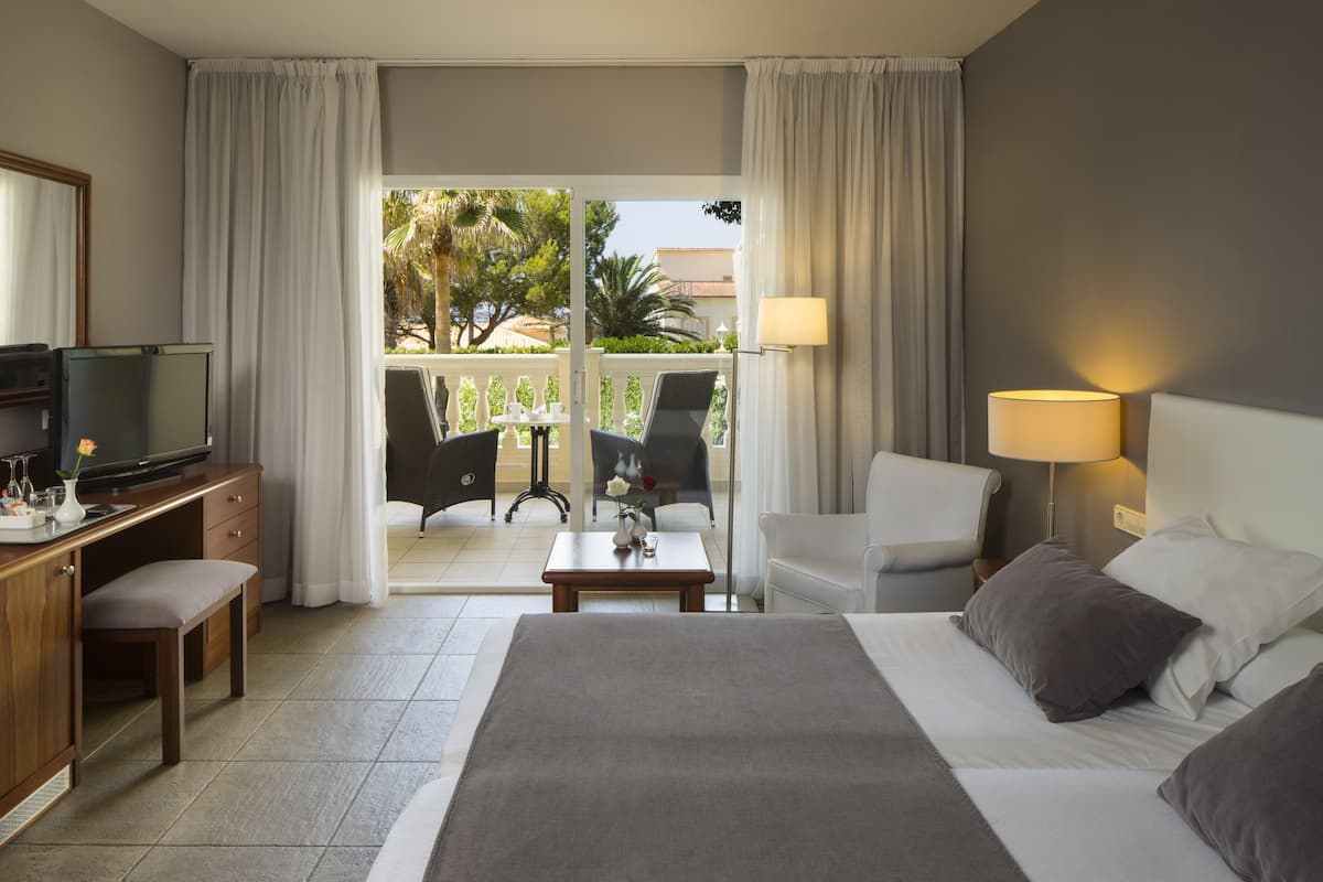 A hotel room with a bed, a desk with a TV, a chair, a lamp, and a balcony with a view of palm trees and buildings.