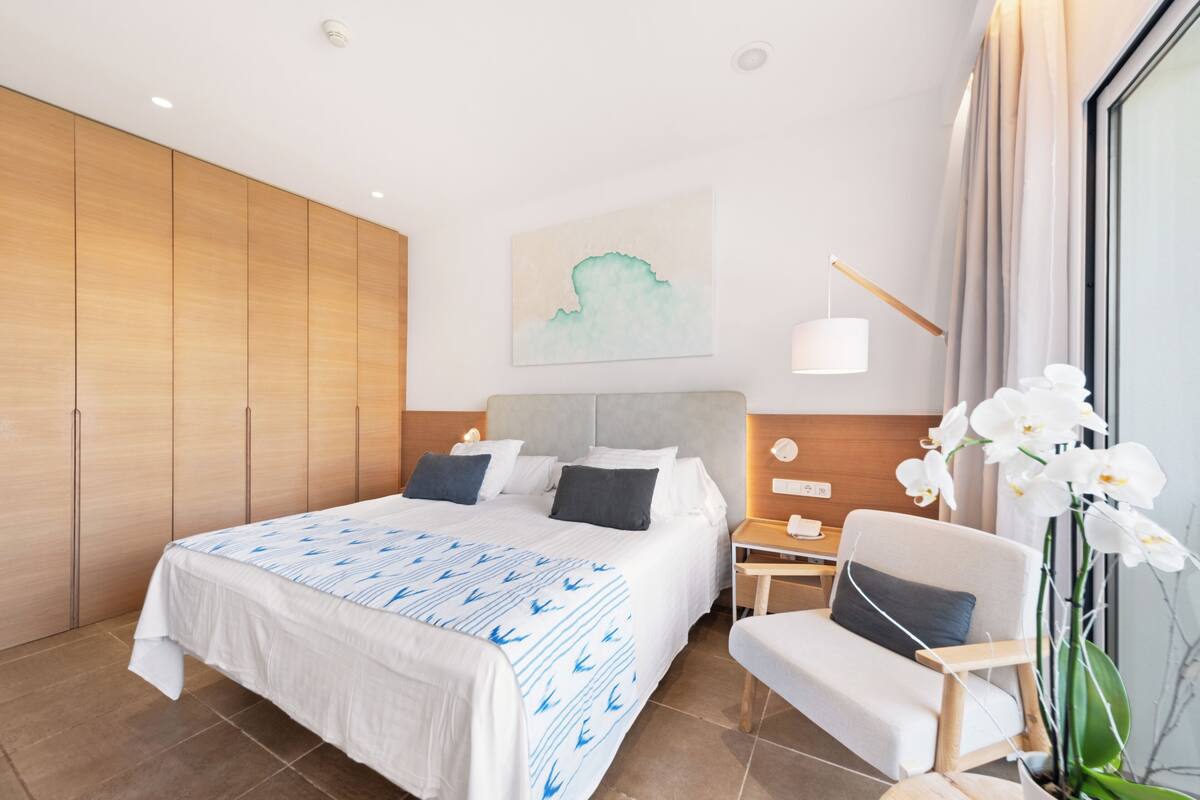 A modern hotel room with a large bed, a wooden wardrobe, a nightstand, a desk, a chair, and a potted orchid.