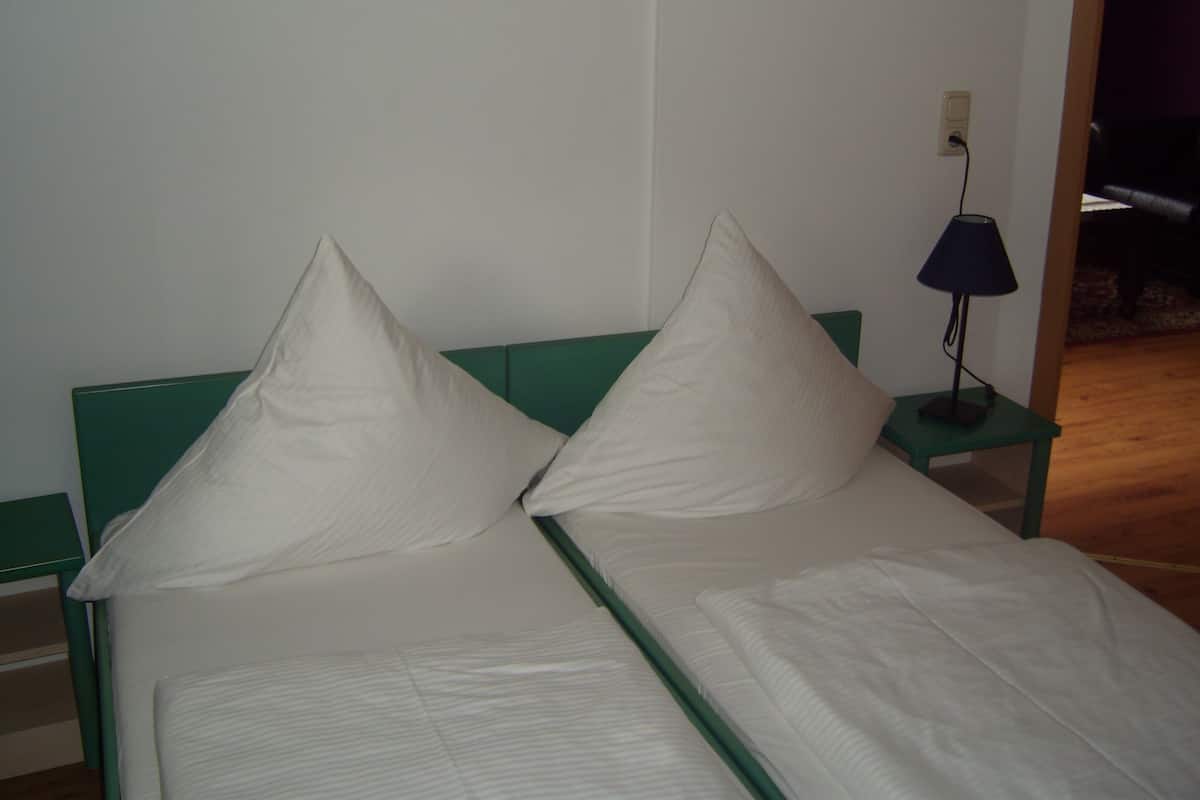 Ein Doppelbett mit zwei Kissen und einem grünen Kopfteil. Neben dem Bett steht ein kleiner Tisch mit einer Lampe.