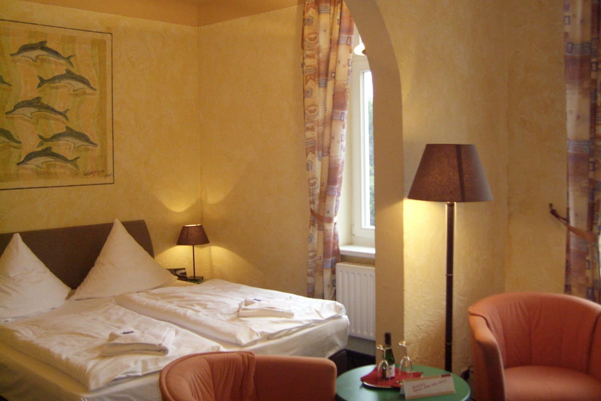 Ein Hotelzimmer mit einem Bett, zwei Stühlen, einem Tisch mit Flasche und Gläsern, einer Lampe und einem Fenster mit Vorhängen.