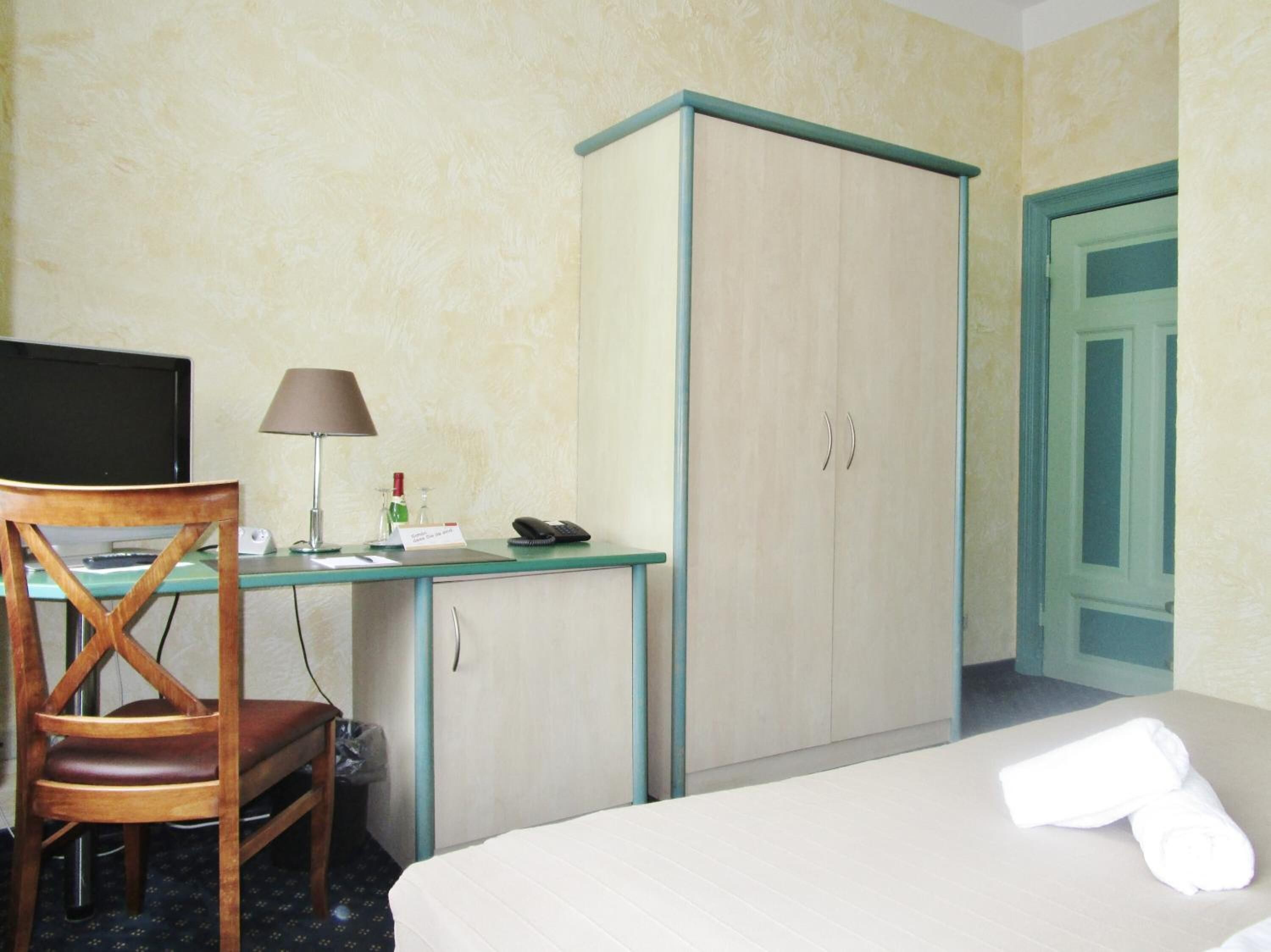 A hotel room with a bed, a desk with a computer, a chair, a wardrobe, and a bathroom door.