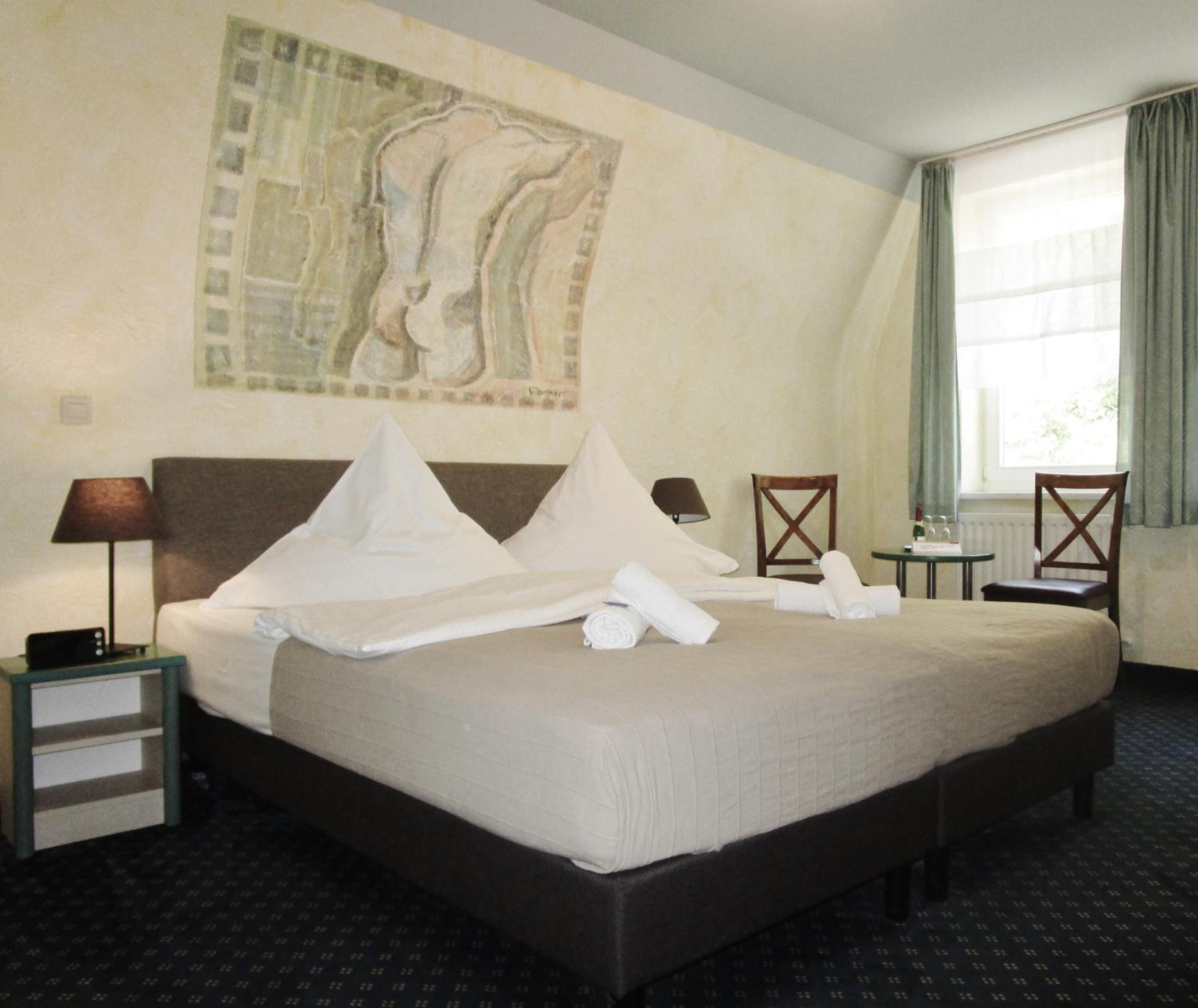 Habitación de hotel con una cama grande, una mesita de noche, una lámpara, una silla, una pequeña mesa y una ventana con cortinas.