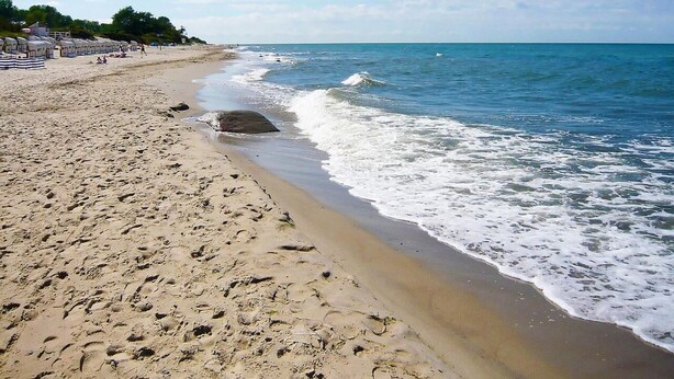 Una playa arenosa con un promontorio rocoso, cielo azul despejado y mar tranquilo.
