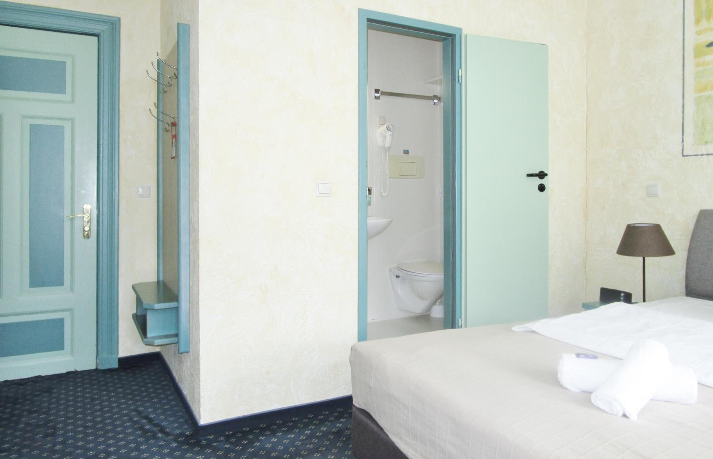 A hotel room with a bed, a bathroom with a toilet and shower, and a small shelf with toiletries.