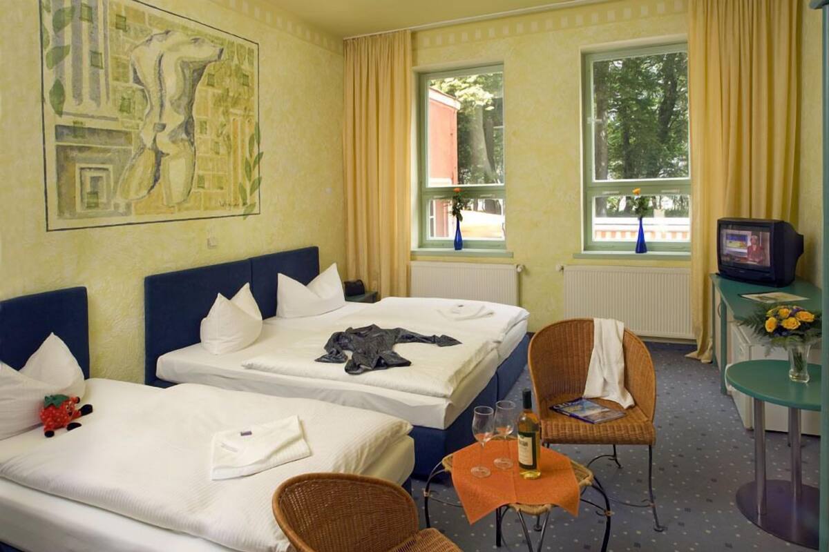 Ein Hotelzimmer mit zwei Betten, einem Fernseher, einem kleinen Tisch mit einer Vase und einem Fenster mit Vorhängen.