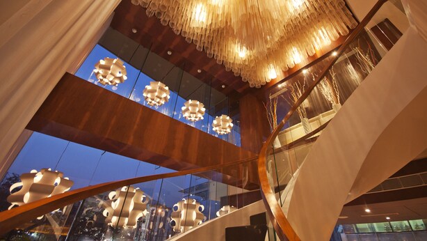 Un interior moderno con una gran araña ornamentada, barandales de vidrio y múltiples luminarias.