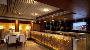 Un área de bar moderna con un revoque de mosaico, una zona de estar con mesas y sillas, y una pantalla de televisión que muestra un paisaje urbano.