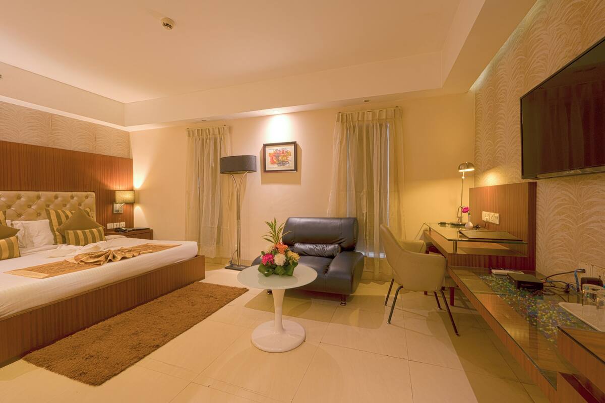 Quarto de hotel com cama, sofá, pequena mesa com arranjo de flores, escrivaninha com computador e televisão na parede.