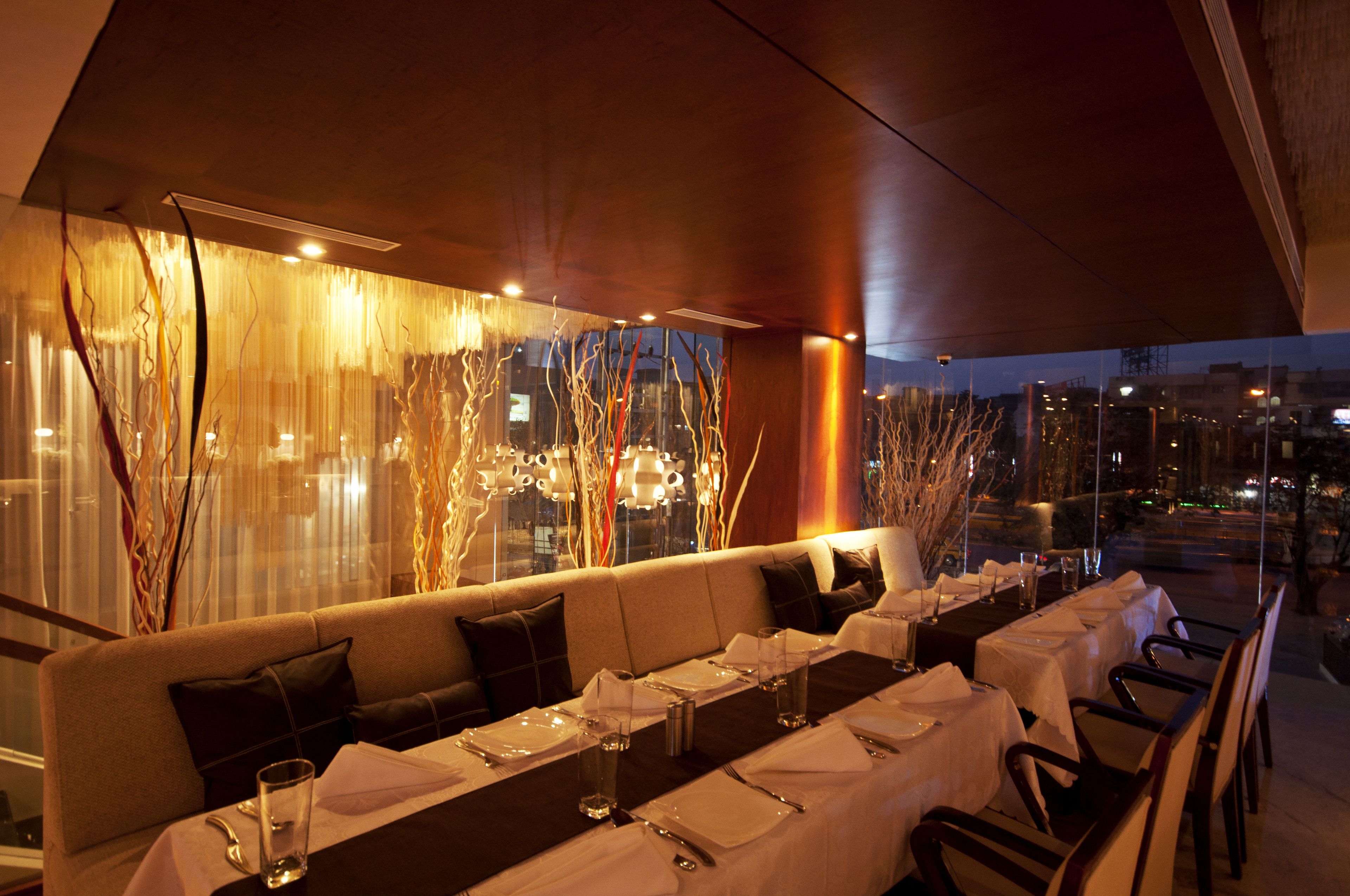A restaurant with a long table set for dining, a cushioned bench, and a view of the cityscape at night.