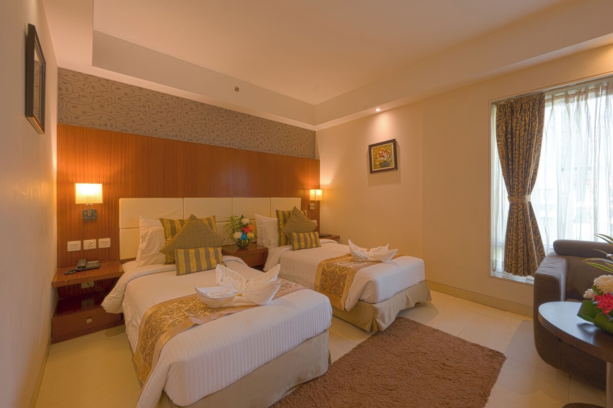 A hotel room with two beds, a sitting area, a window with curtains, and a small table with flowers.