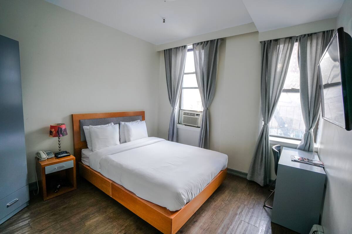 Een hotelkamer met een houten bed, een nachtkastje met een lamp, een bureau en een raam met gordijnen.