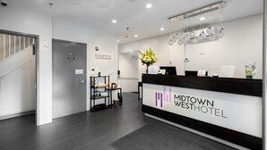 A reception area with a reception desk, a sign for the Midtown West Hotel, a staircase, and a small shelf with items. - Midtown West Hotel (New York)
