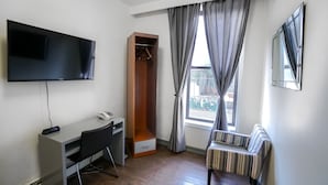 A modern hotel room with a flat-screen TV, a desk with a chair, a wardrobe, a bench, and a large window with curtains. - Midtown West Hotel (New York)