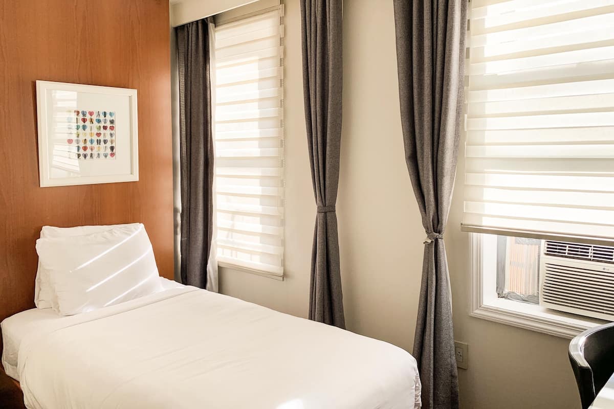 Een hotelkamer met een net opgemaakt bed, een ingelijst schilderij aan de muur en grote ramen met gordijnen.