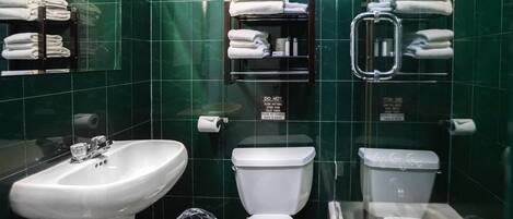 A modern bathroom with green tiles, a sink, two toilets, and a towel rack with folded towels.