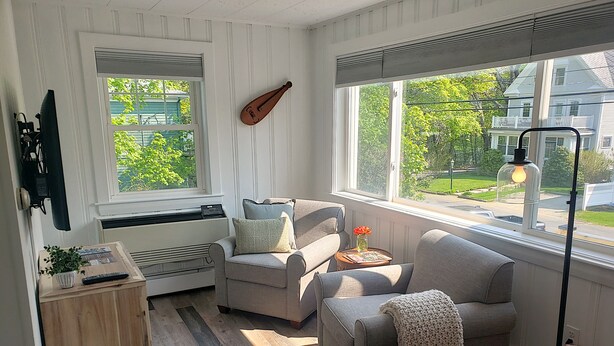 A cozy living room with a grey sofa, a small table with a lamp, and a window with a view of a house and trees. - Little Fig Hotel (Bar Harbor)