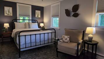A bedroom with a metal bed frame, a beige armchair, and a small table with a lamp. There are two windows with blinds, a ceiling light, and framed artwork on the walls.