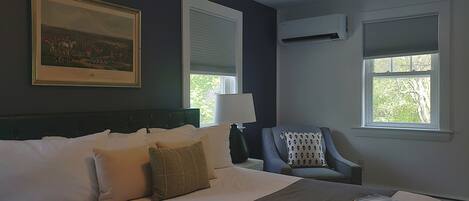 A hotel room with a large bed, a chair, a painting on the wall, and two windows with blinds.