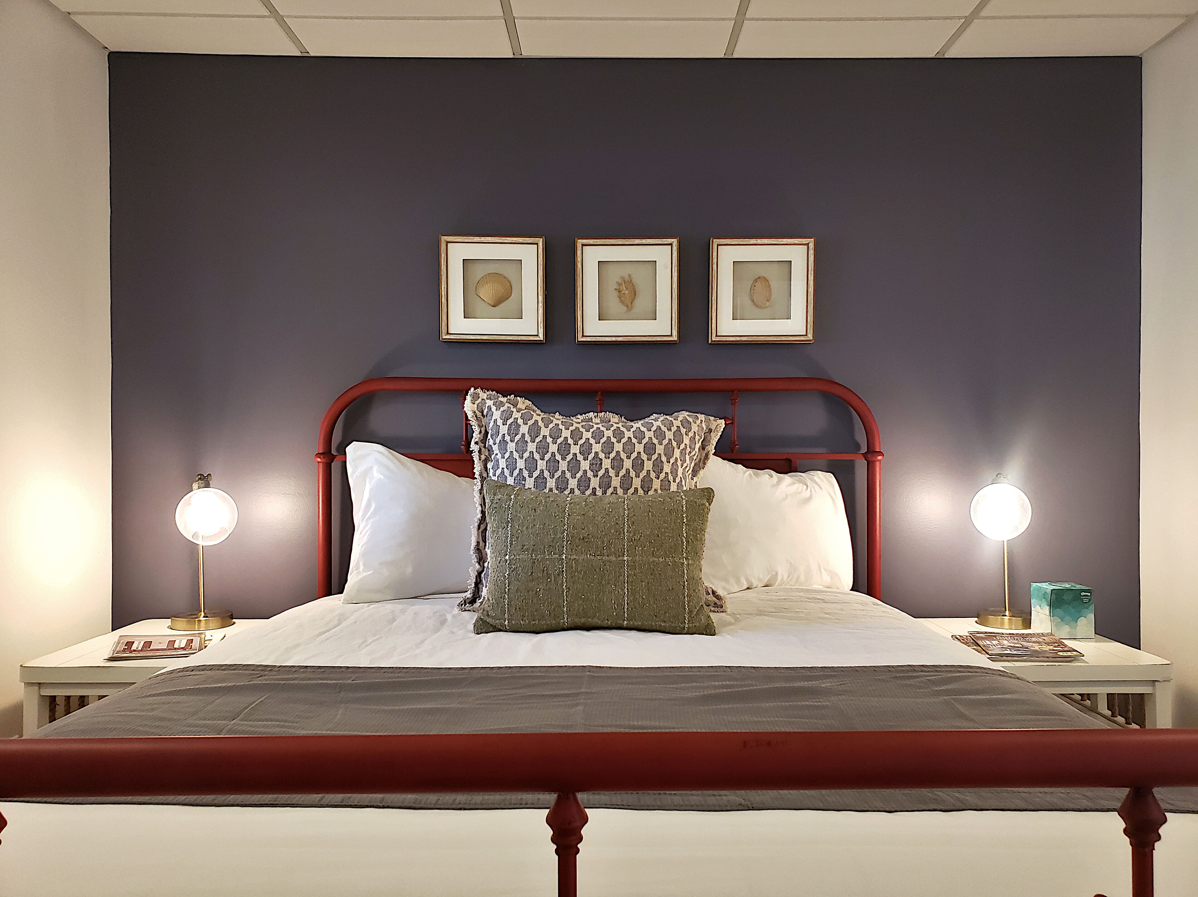 A neatly made bed with a red metal frame, white bedding, and multiple pillows. There are two bedside tables with lamps, and three framed pictures on the wall above the bed.