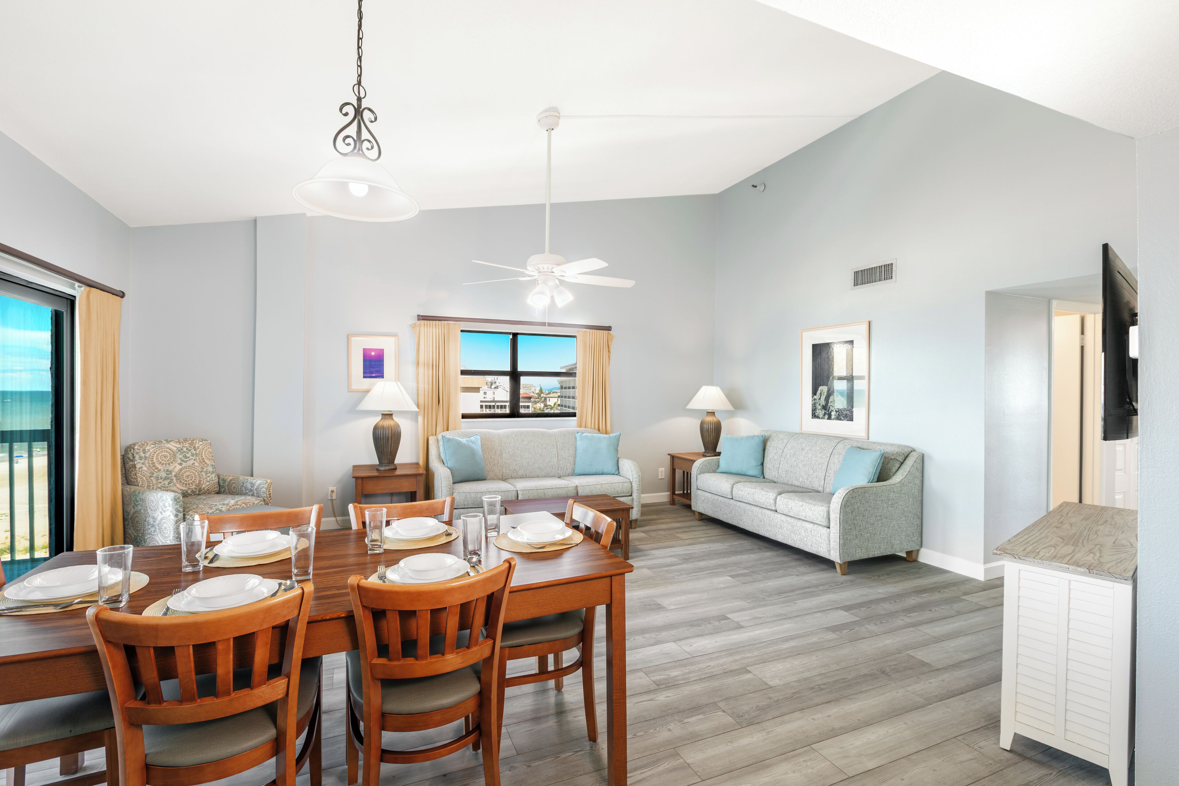 A dining area with a wooden table set for four, a sofa, and a television in a room with light blue walls and a view through a window.