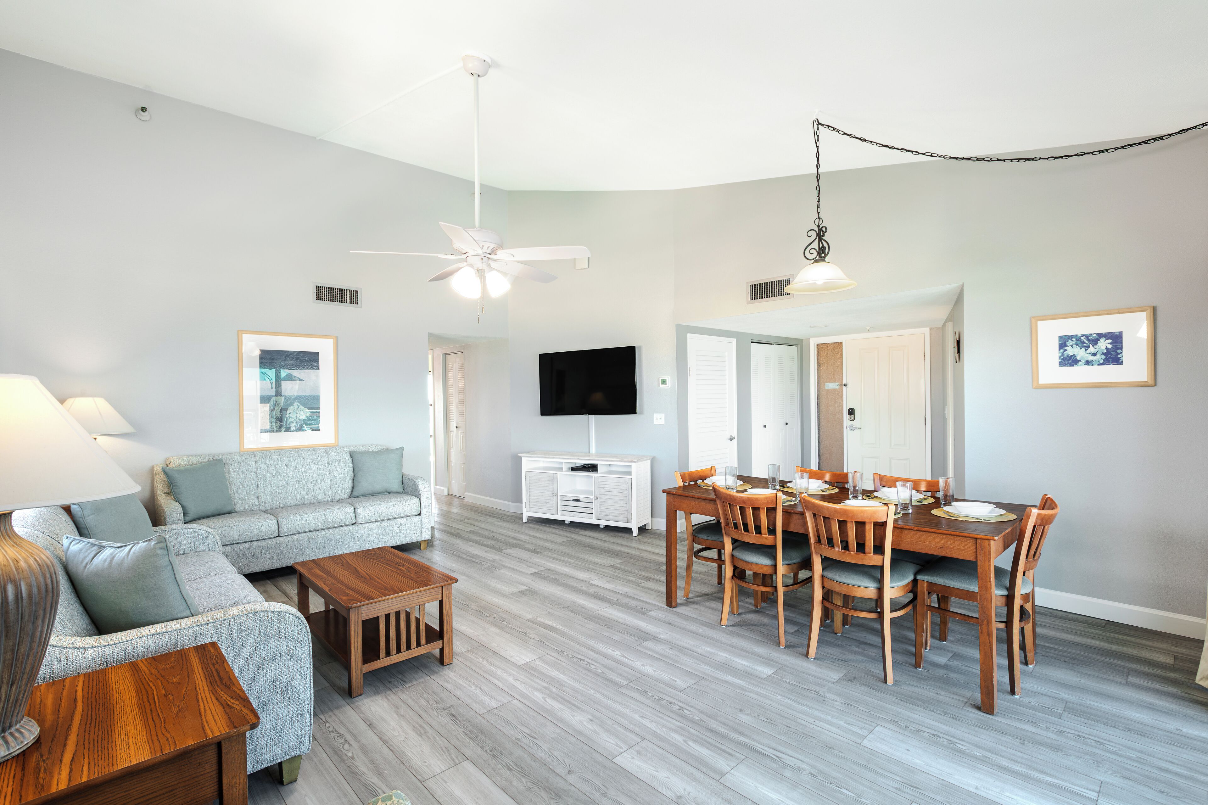 A modern living room with a grey sofa, wooden coffee table, dining table with chairs, a ceiling fan, and a flat-screen TV mounted on the wall.