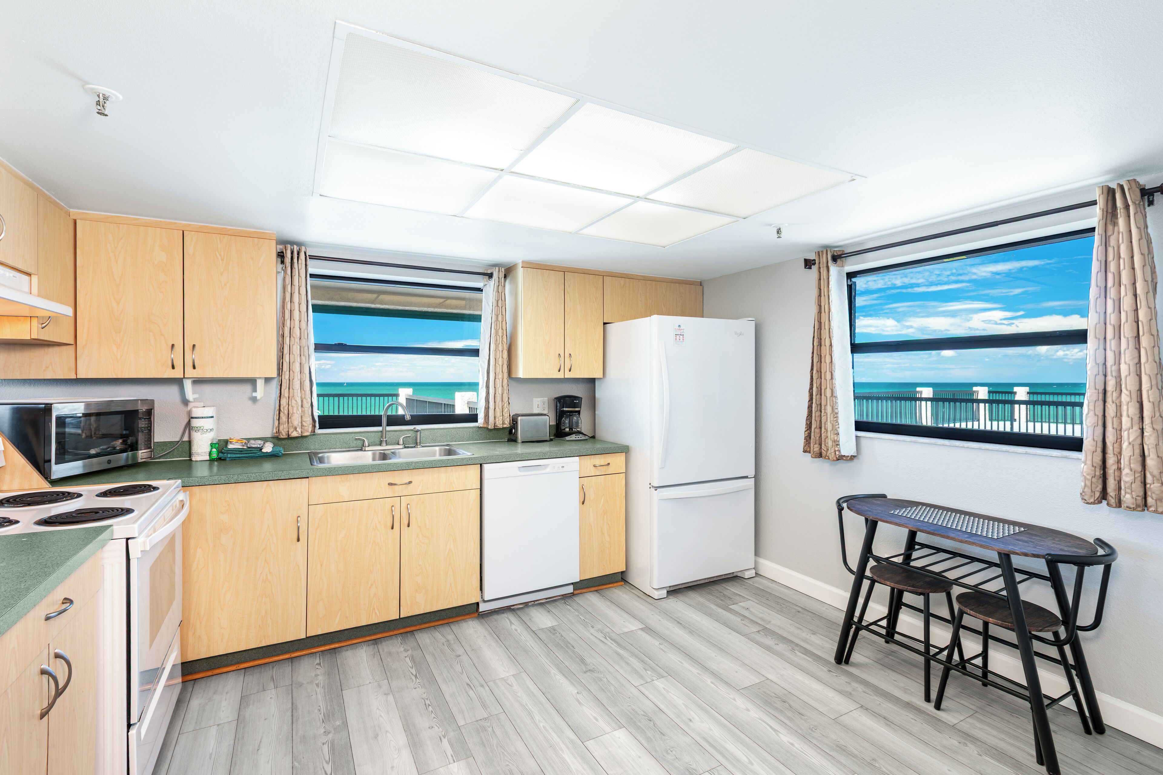 A modern kitchen with wooden cabinets, a white refrigerator, a microwave, and a sink. There is a window with a view of the sea.