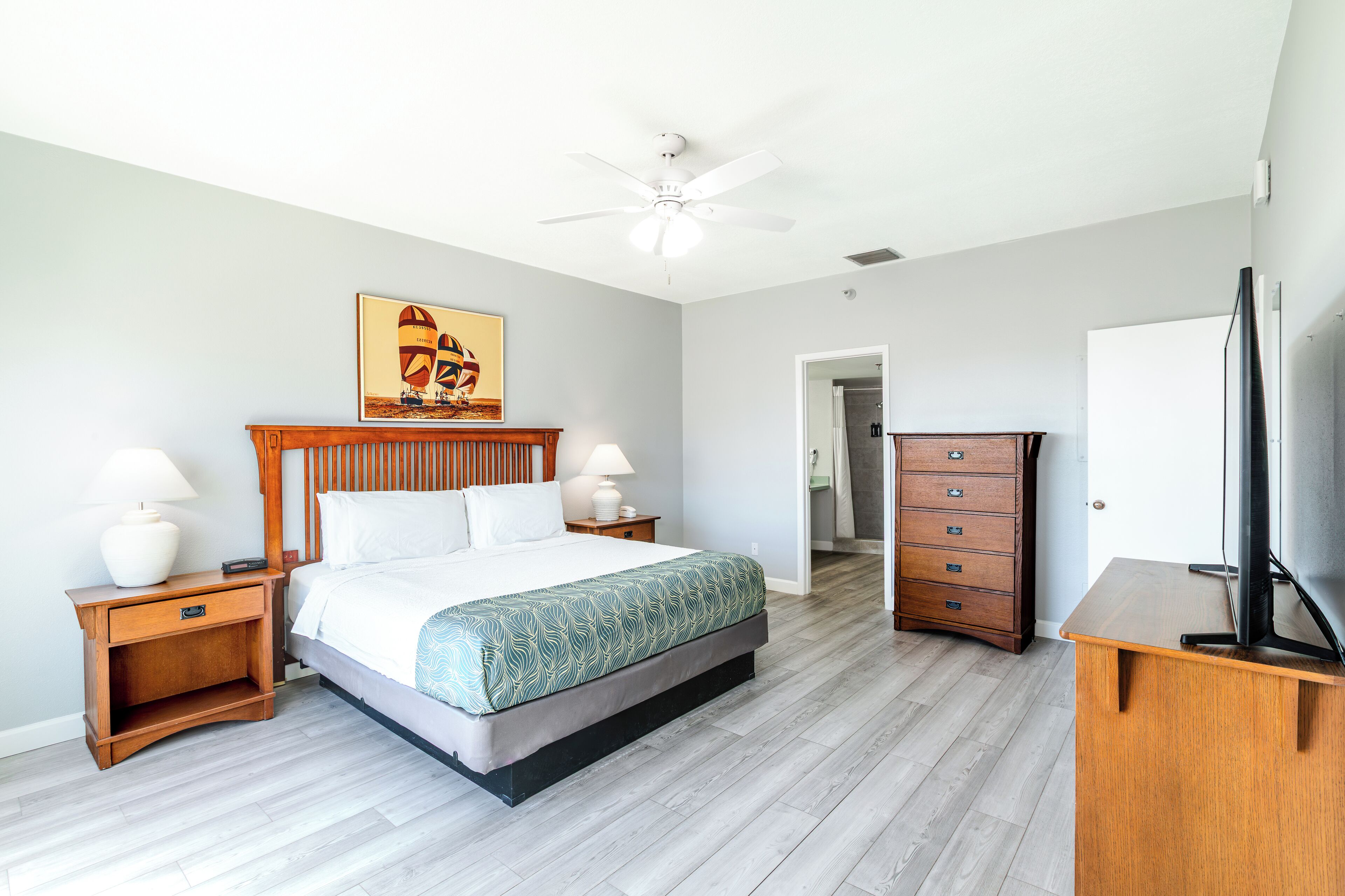 Une chambre d’hôtel comprenant un lit, une table de chevet en bois, une commode, un téléviseur et un ventilateur de plafond.