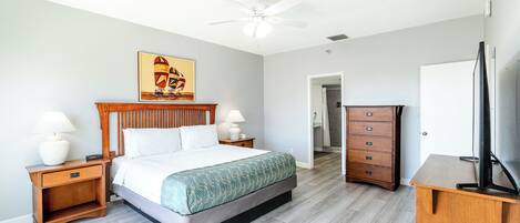 Une chambre d’hôtel comprenant un lit, une table de chevet en bois, une commode, un téléviseur et un ventilateur de plafond.