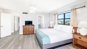Un dormitorio con cama, cómoda de madera, televisor, lámparas de noche y ventana con cortinas.