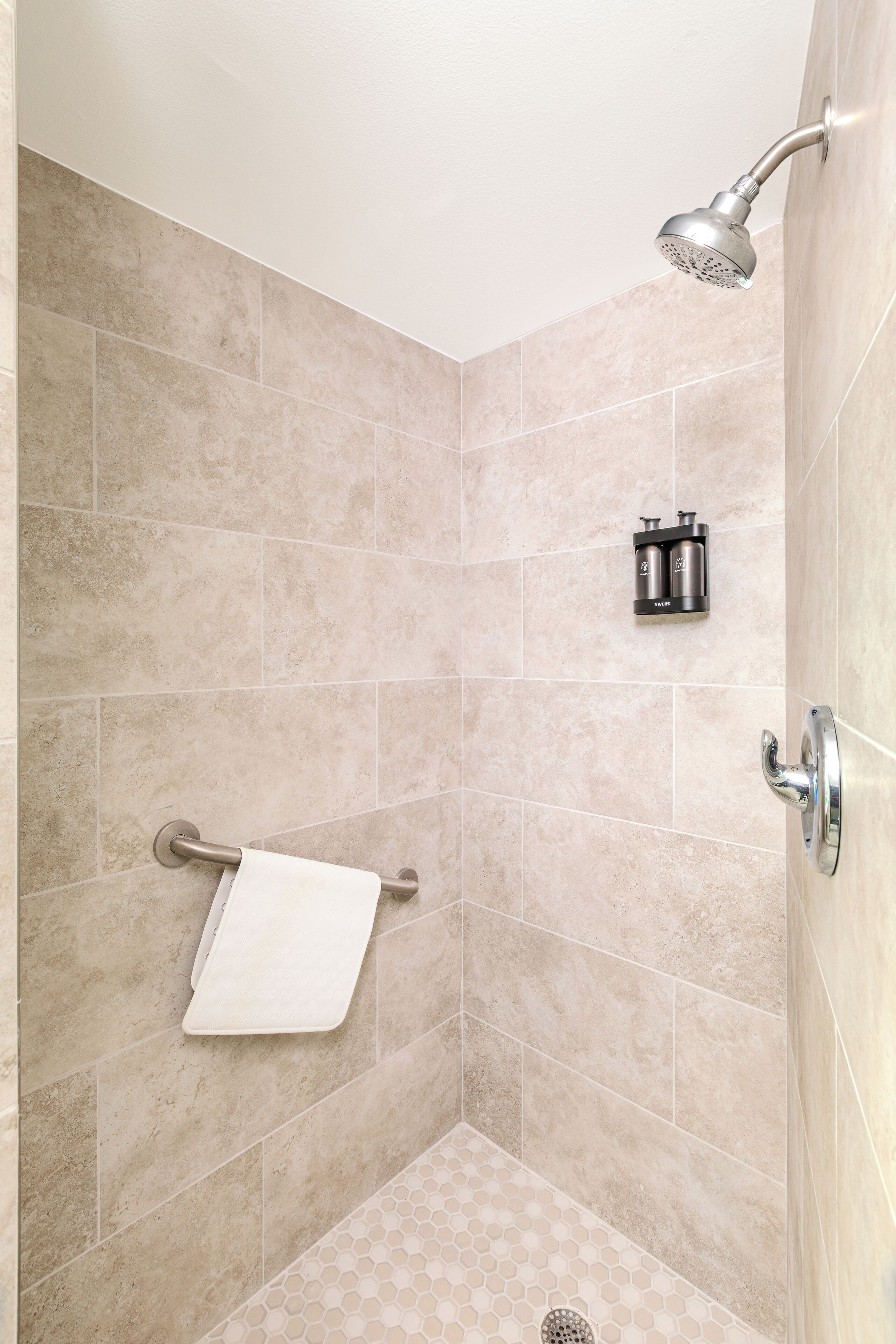 Ducha con cabezal de ducha de mano, toallero y dispensador de jabón adherido a la pared en un baño con azulejos.