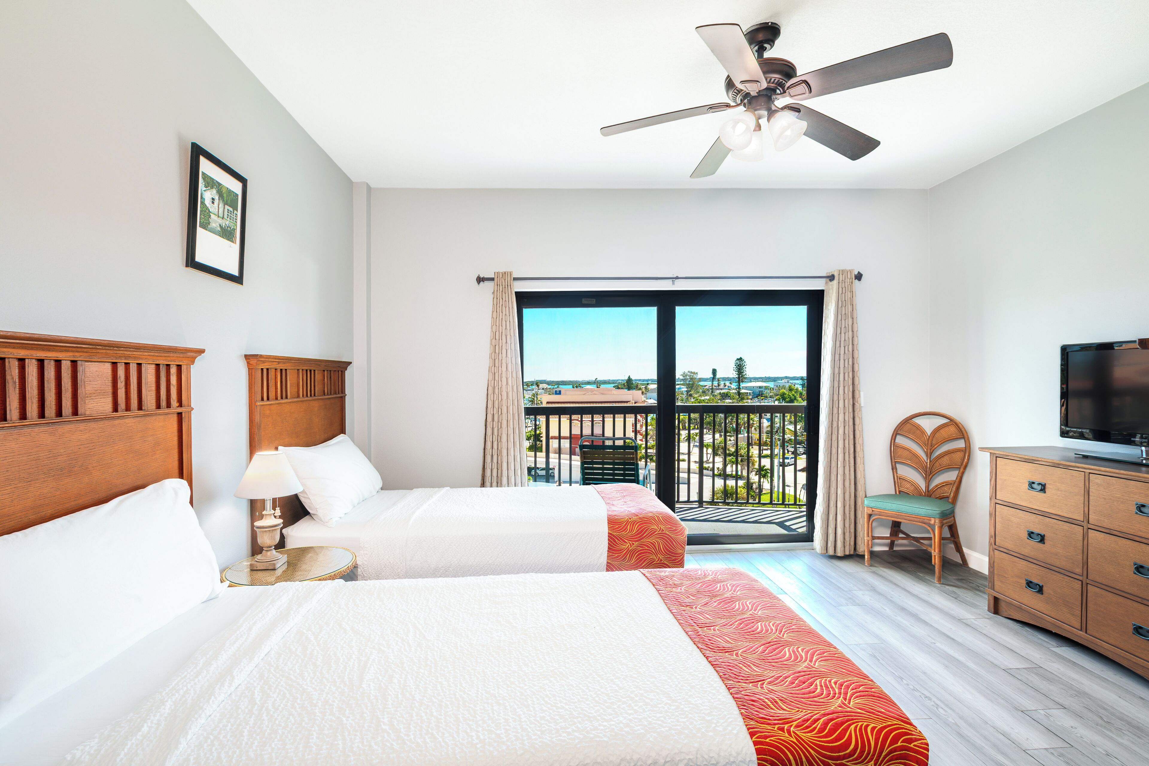 Habitación de hotel con dos camas, televisor, ventilador de techo y balcón con vistas.
