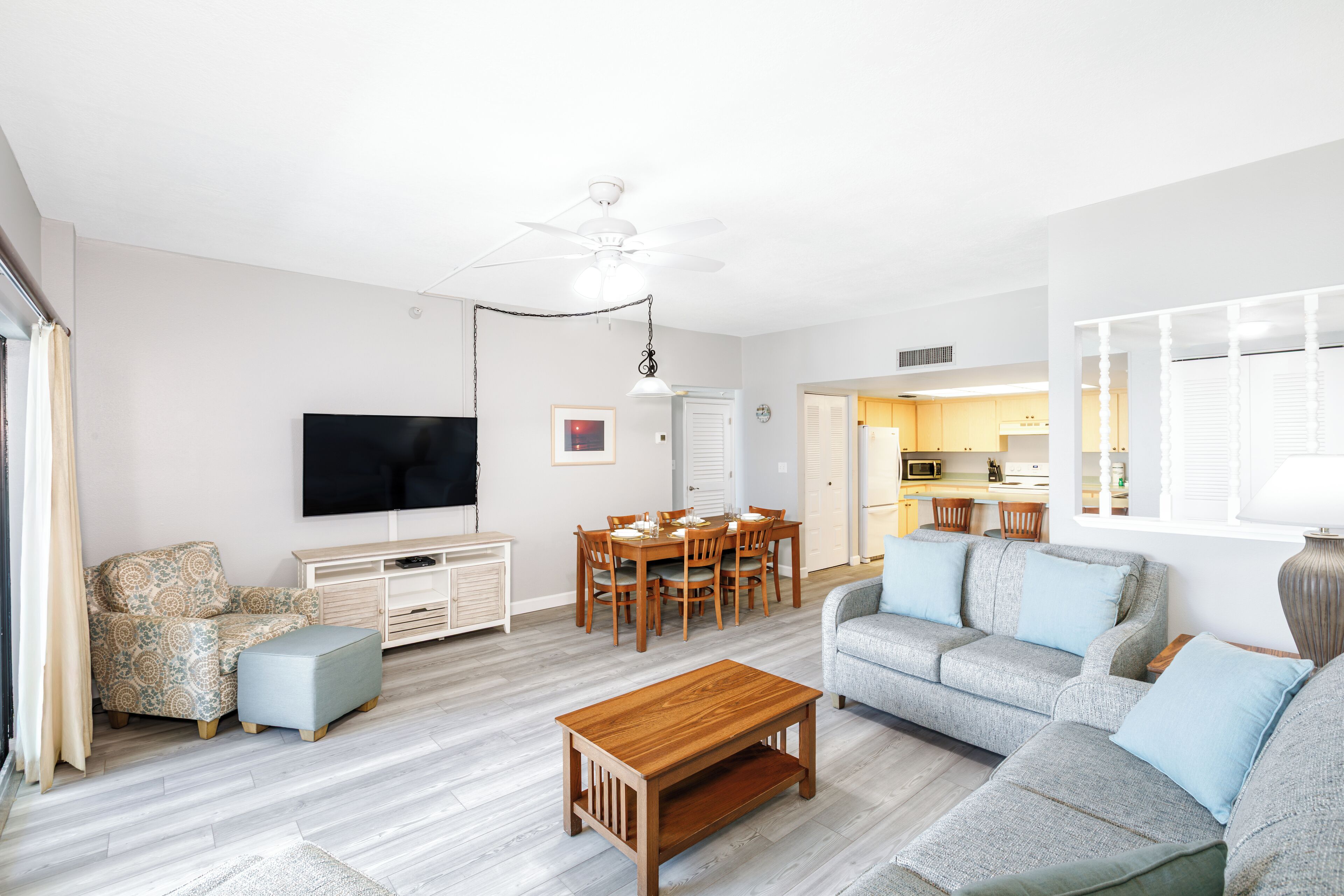 A spacious living room with a flat-screen TV, a dining area with a wooden table and chairs, a grey sofa, and a wooden coffee table.