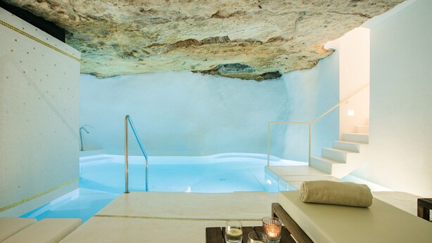 Un cuarto de spa con techo de piedra, una bañera de hidromasaje, un banco de madera y una mesa con velas.