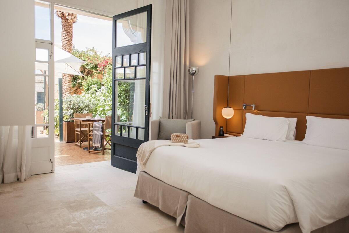 A hotel room with a bed, bedside table, lamp, and a view of a garden through an open door.