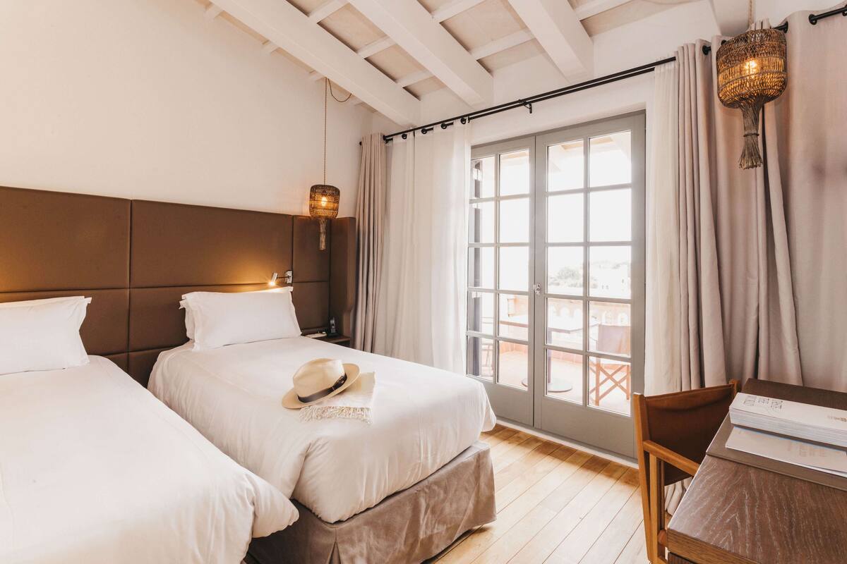 A hotel room with two beds, a wooden floor, a balcony with curtains, and a hanging light fixture.