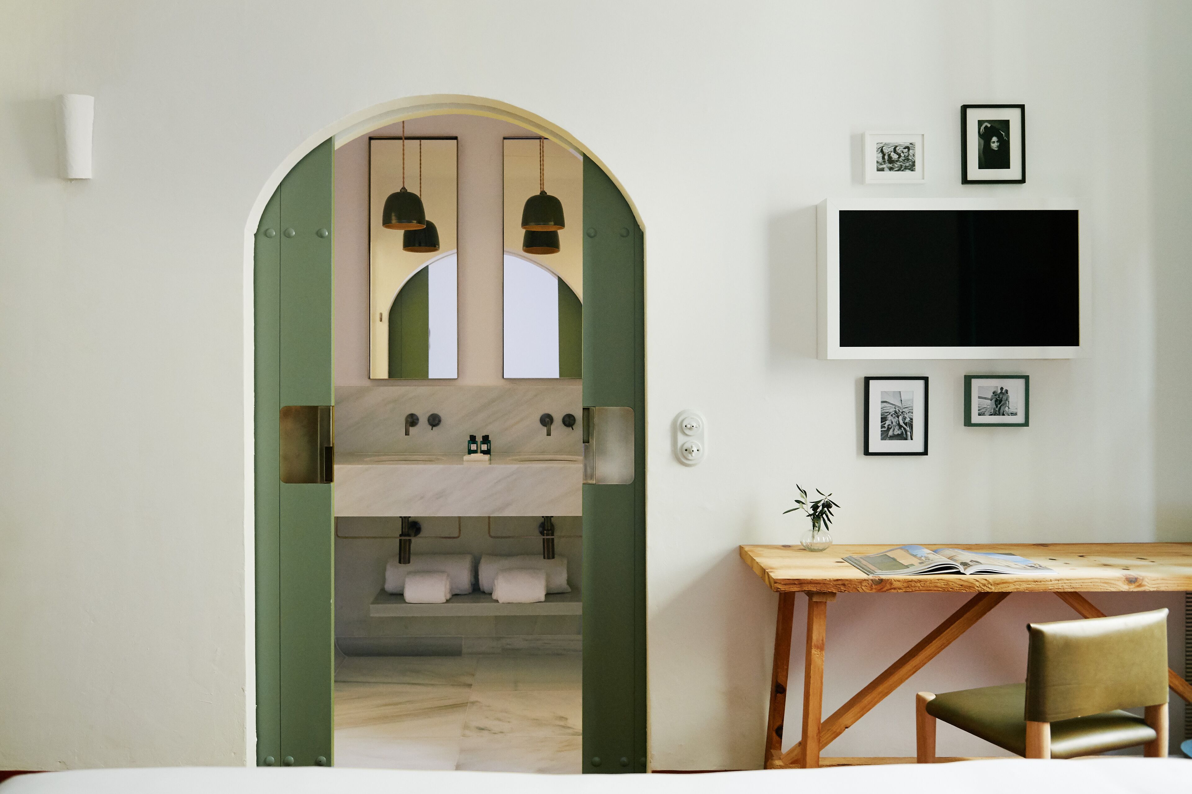 A modern bathroom with a green archway, a wooden desk with a chair, and a flat-screen TV mounted on the wall.