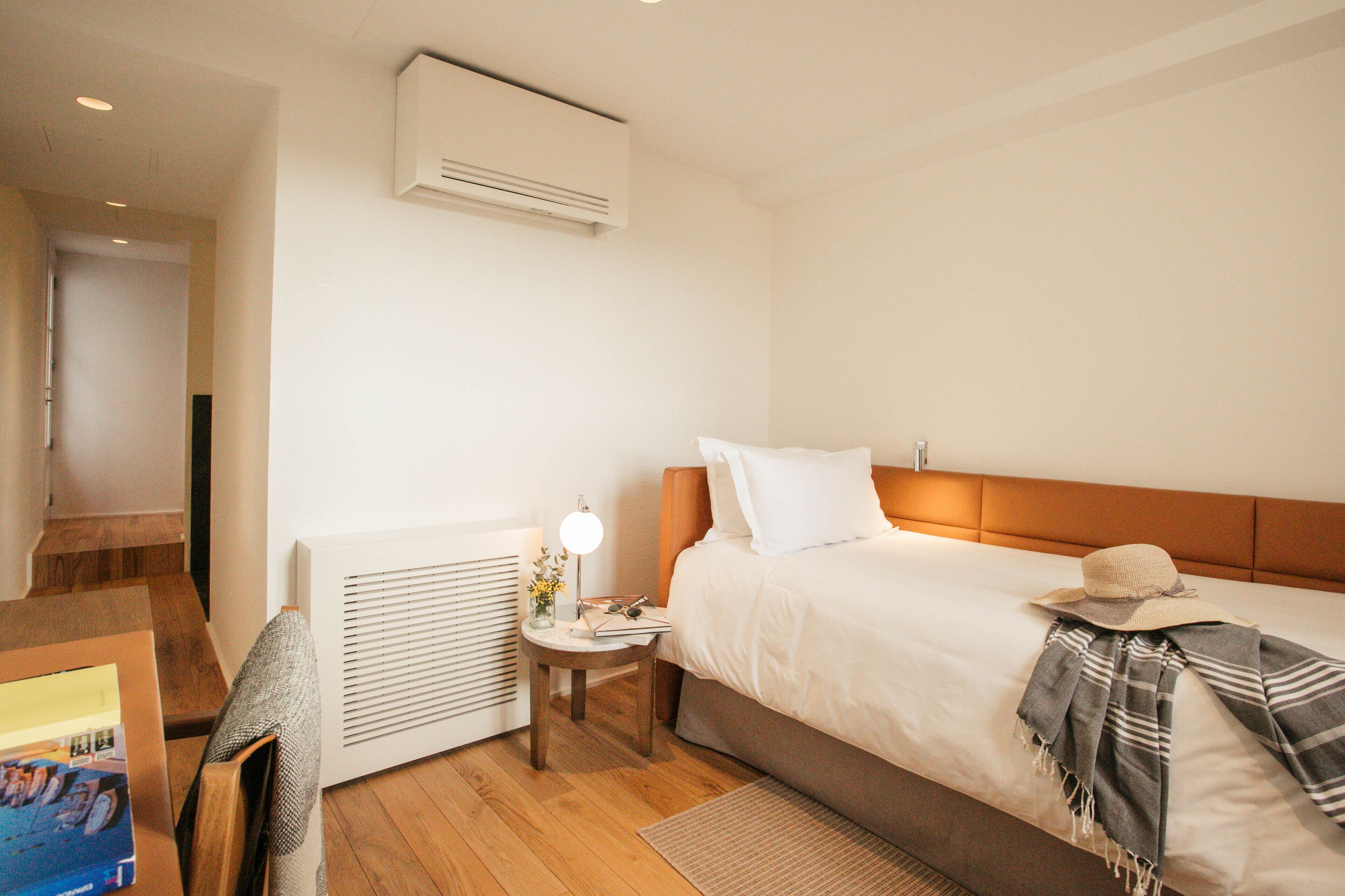A hotel room with a wooden floor, a bed with white bedding, a nightstand with a lamp, and an air conditioning unit on the wall.