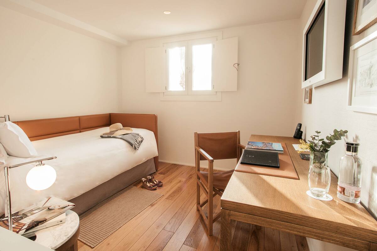A small, well-lit bedroom with a single bed, a wooden desk, a chair, a small table, a lamp, a vase with flowers, and a framed picture on the wall.