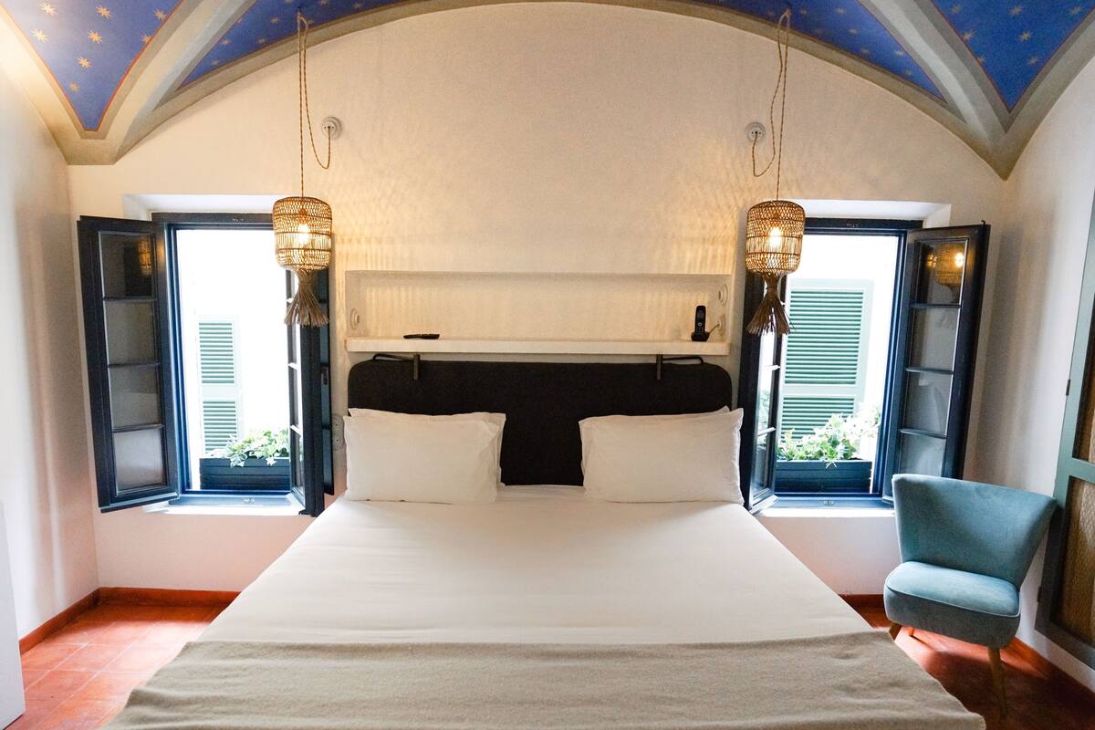A large bed with a dark headboard, two bedside lamps, and a view of greenery through open windows.