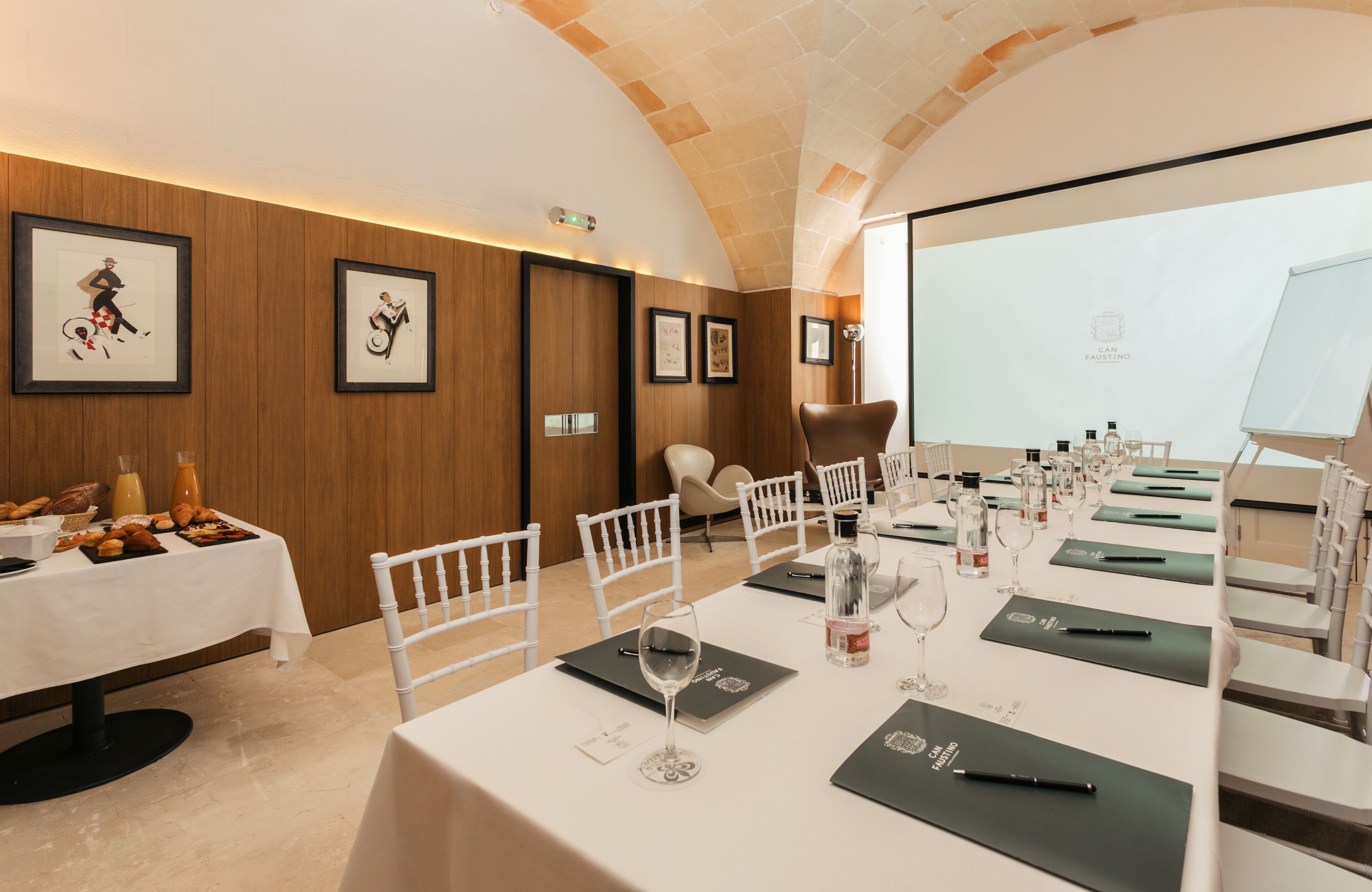 Un salón de conferencias con una larga mesa preparada para una reunión, obras de arte enmarcadas en las paredes y una pantalla de proyector.