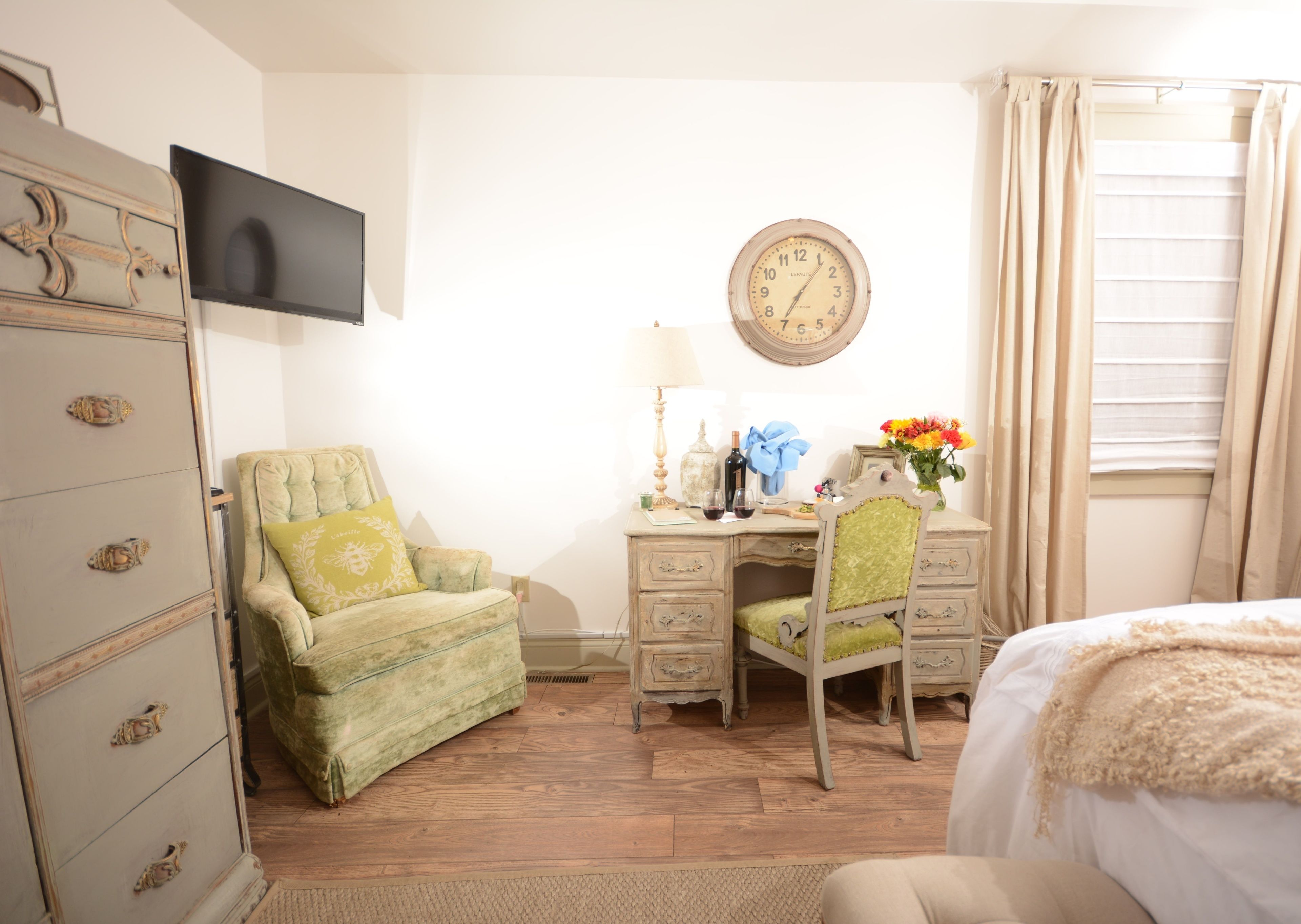 A room with a bed, a desk with a chair, a dresser, a television, a clock, and a window with curtains.