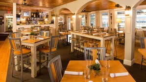Un restaurante con mesas y sillas de madera, una zona de bar y grandes ventanales.