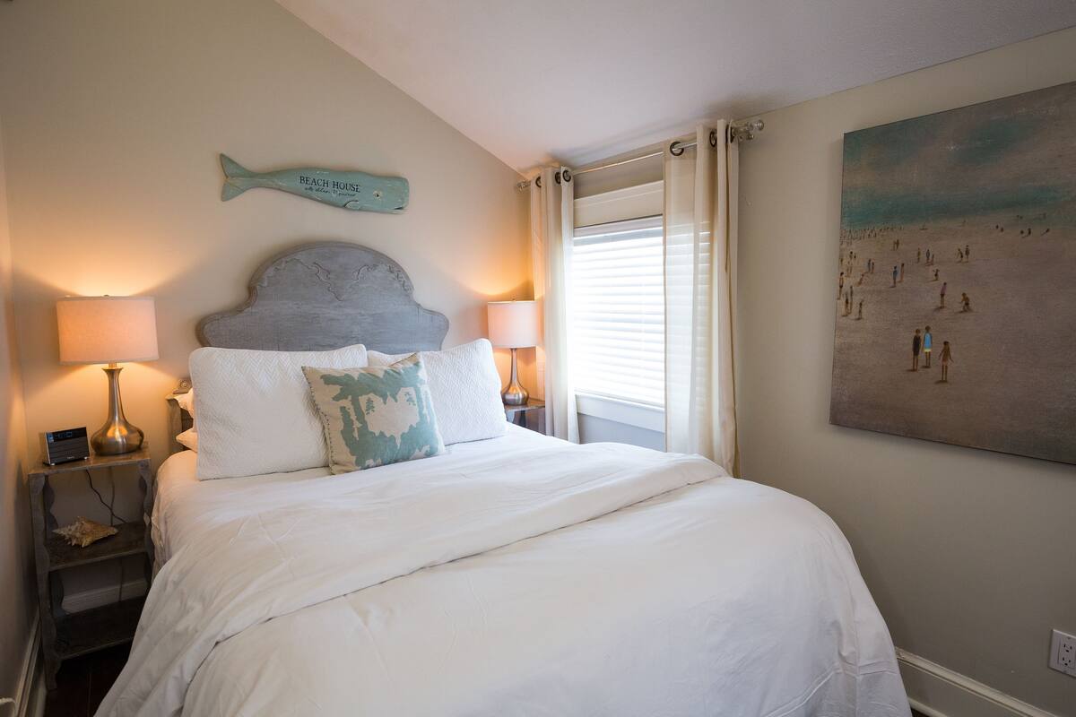 A bedroom with a large bed, two bedside lamps, a wall-mounted fish decoration, and a beach-themed painting.