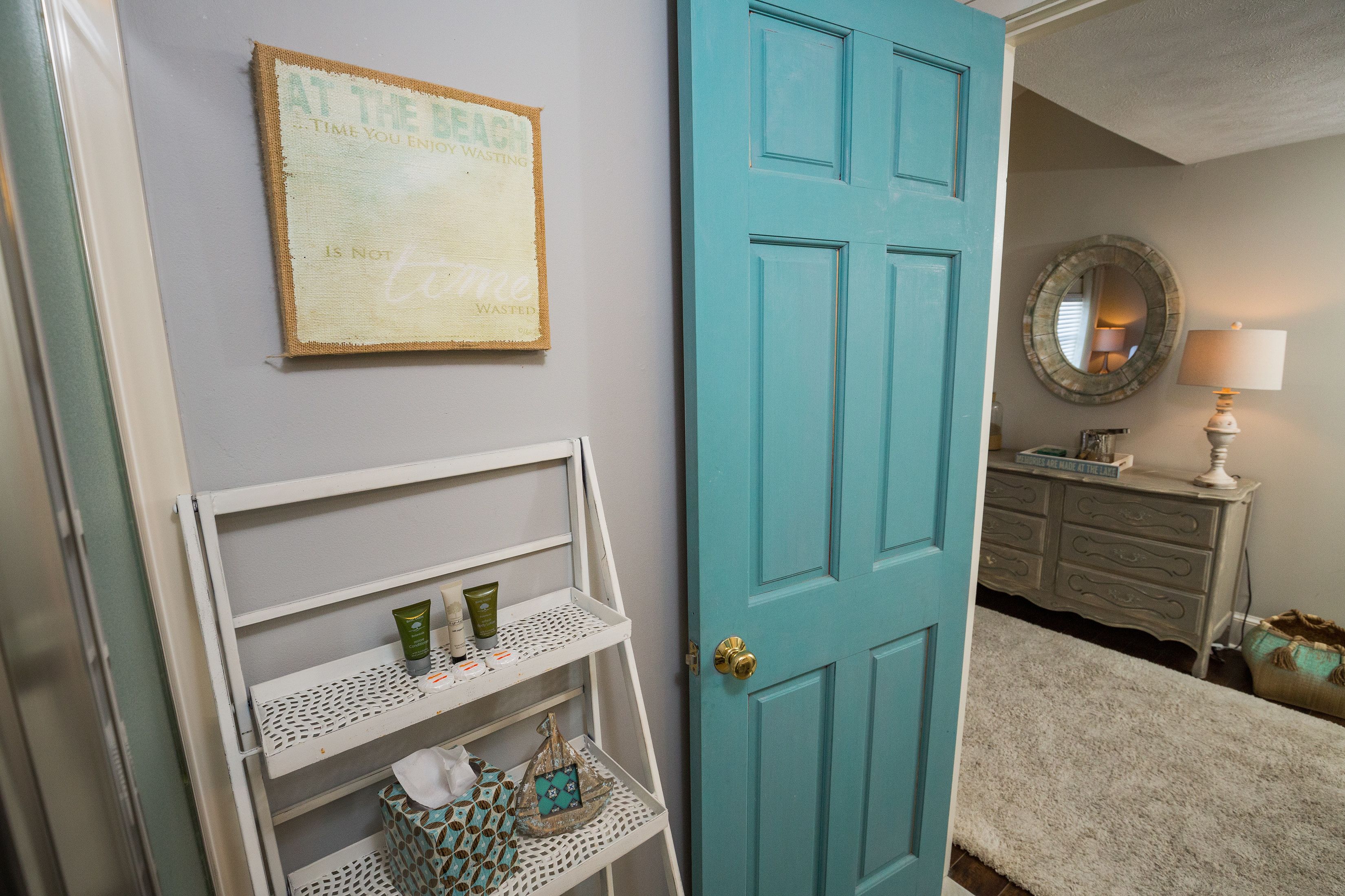 Une salle de bain avec une porte turquoise, une étagère contenant des articles de toilette, un miroir et une lampe.