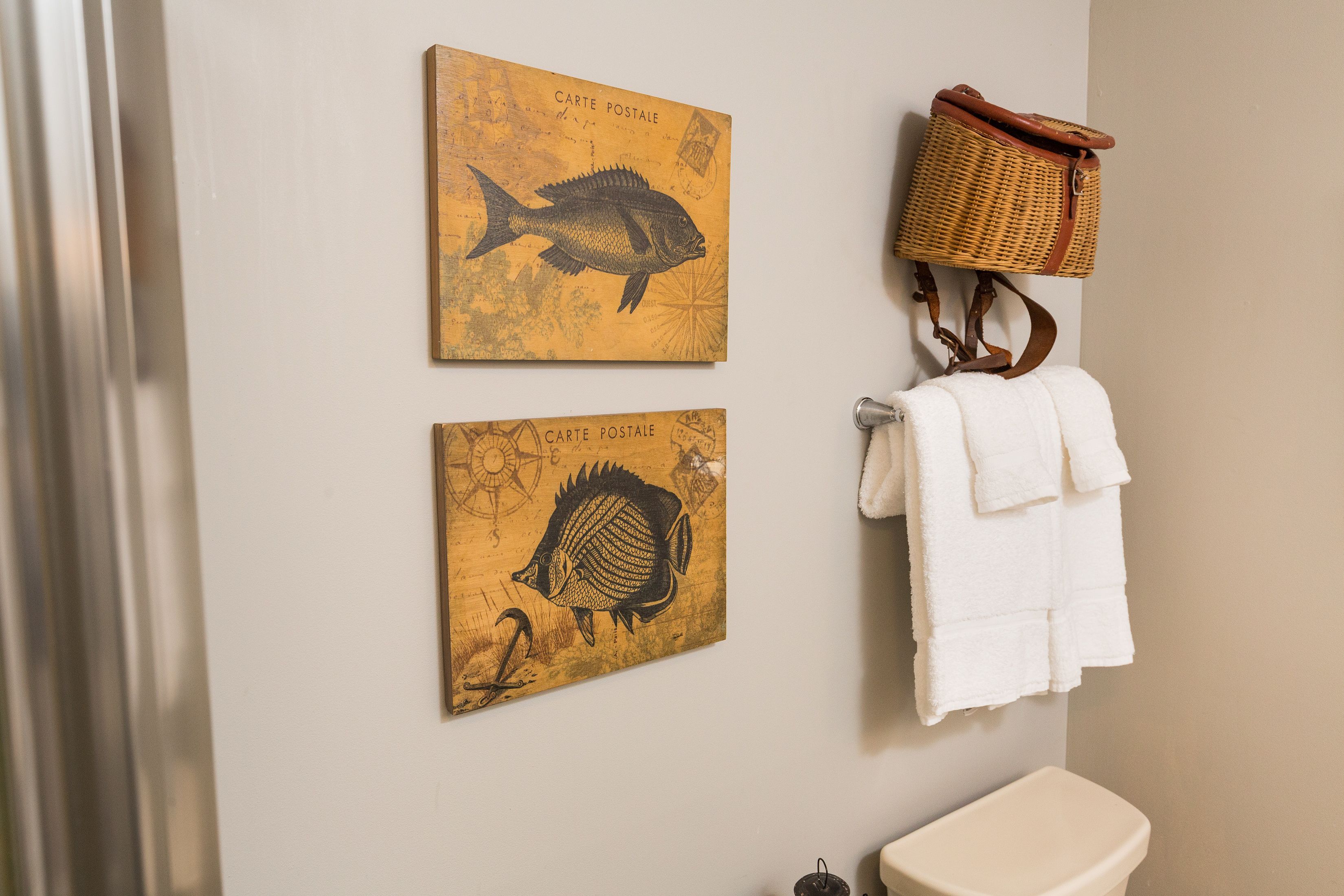 Un baño con dos obras de arte colgadas en la pared que representan peces, una cesta de mimbre y una toalla blanca colgada en un perchero.