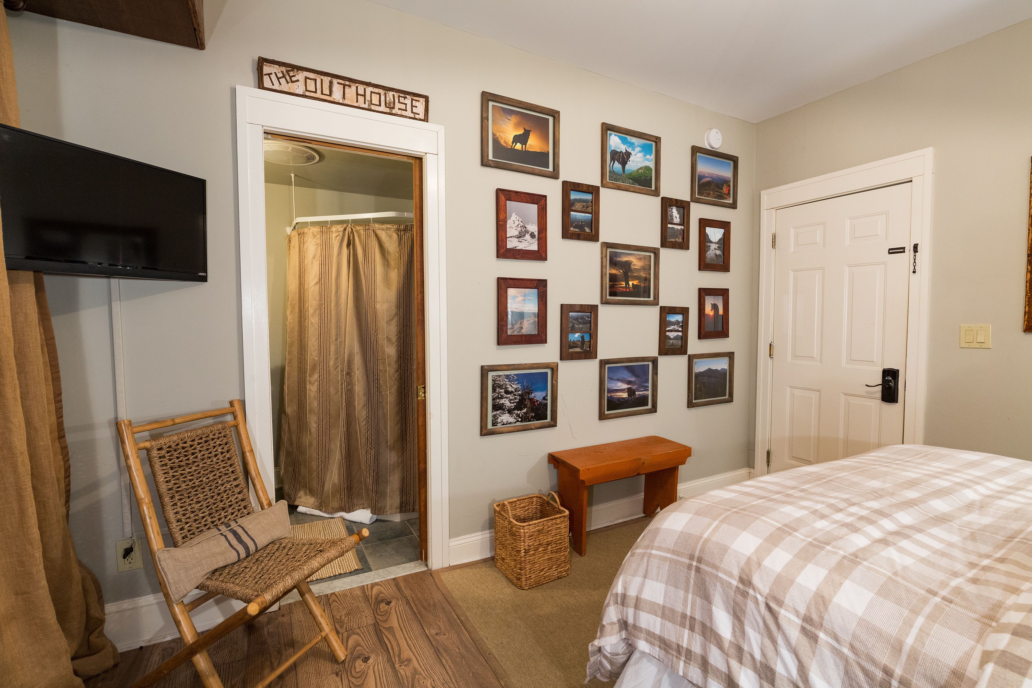 A bedroom with a bed, a chair, a bench, a wall with framed pictures, a door, and a television.