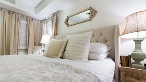 Una cama bien hecha con una colcha de estampado floral, un espejo grande encima y una mesita de noche con libros y una lámpara.