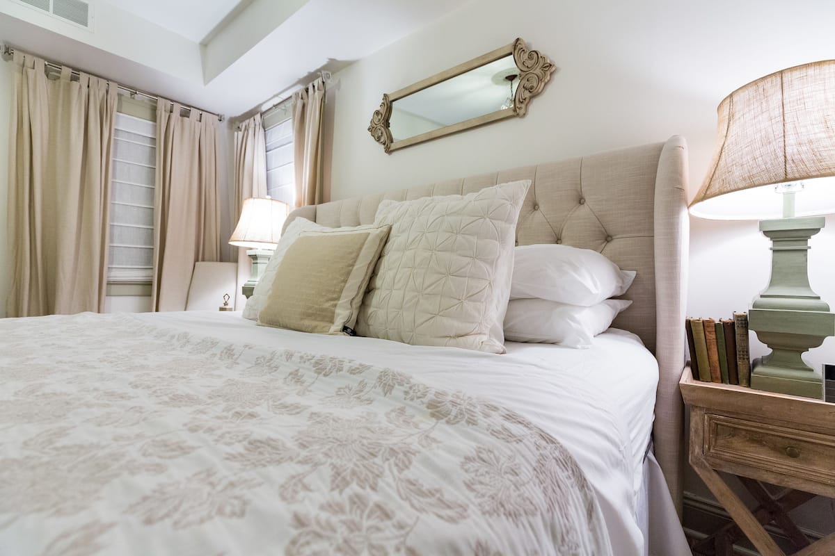 A neatly made bed with a floral-patterned bedspread, a large mirror above it, and a bedside table with books and a lamp.