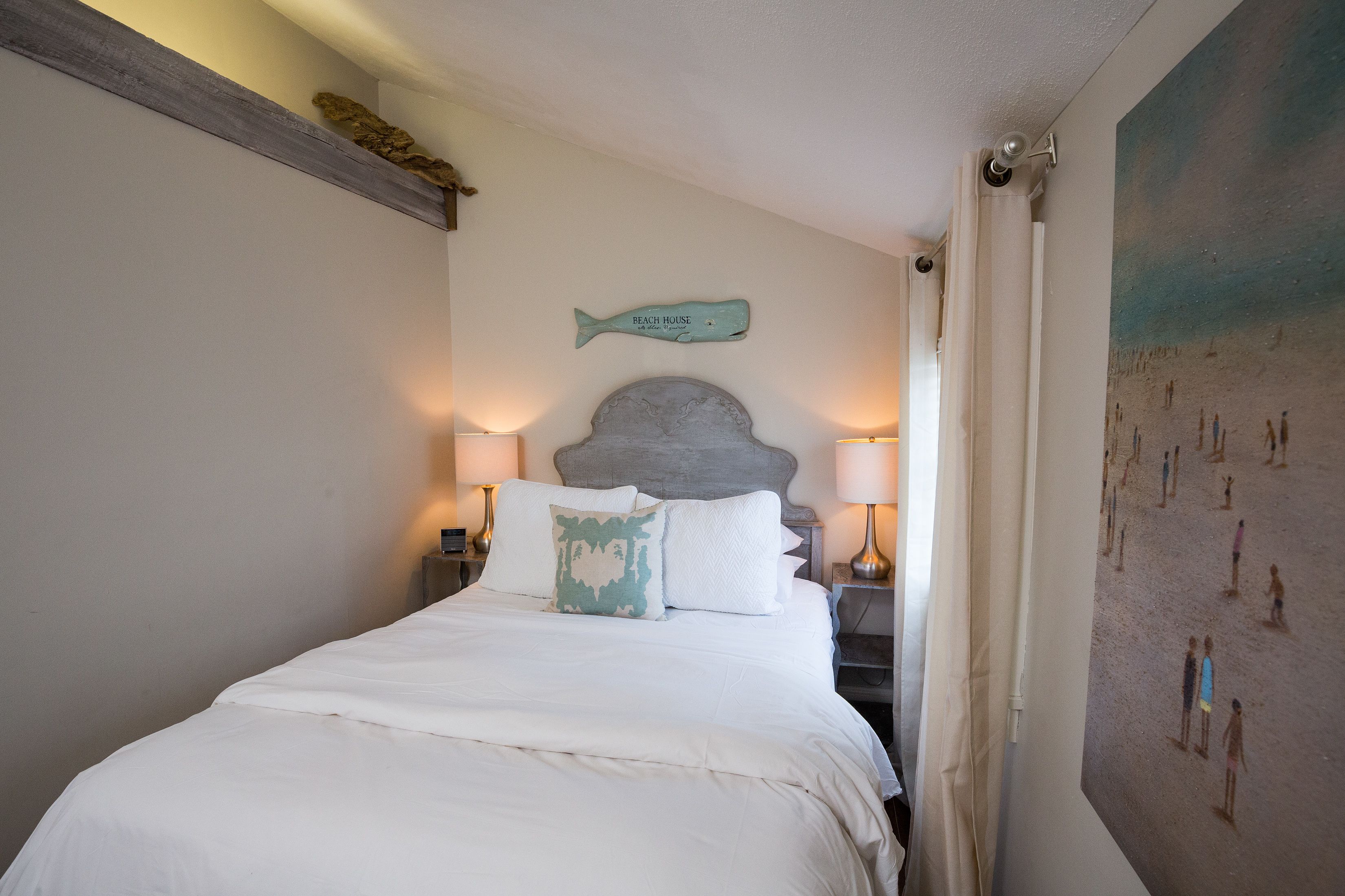 A bedroom with a bed, two bedside lamps, a wall-mounted fish decoration, and a beach-themed painting.